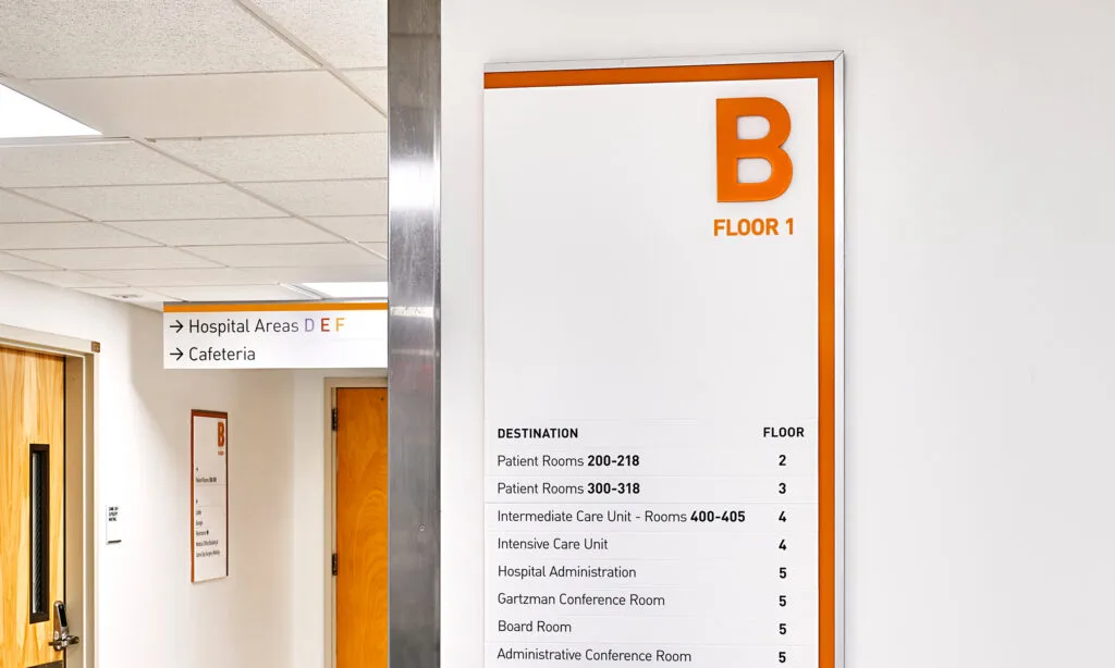 Building directory sign for Floor 1 in Section B of a Hospital, overhead directional sign in background.