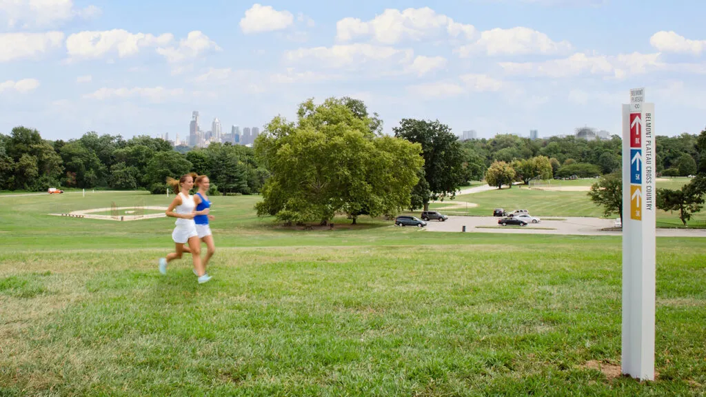 Two runners passing Belmont Plateau pillar identification sign sitting against Philadelphia skyline