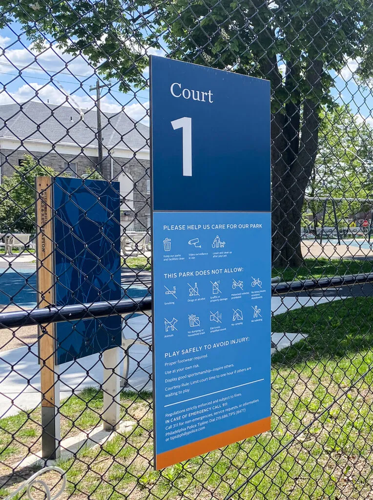 Sign mounted to fence outside basketball court. The sign includes the court number and icons that represent the park rules