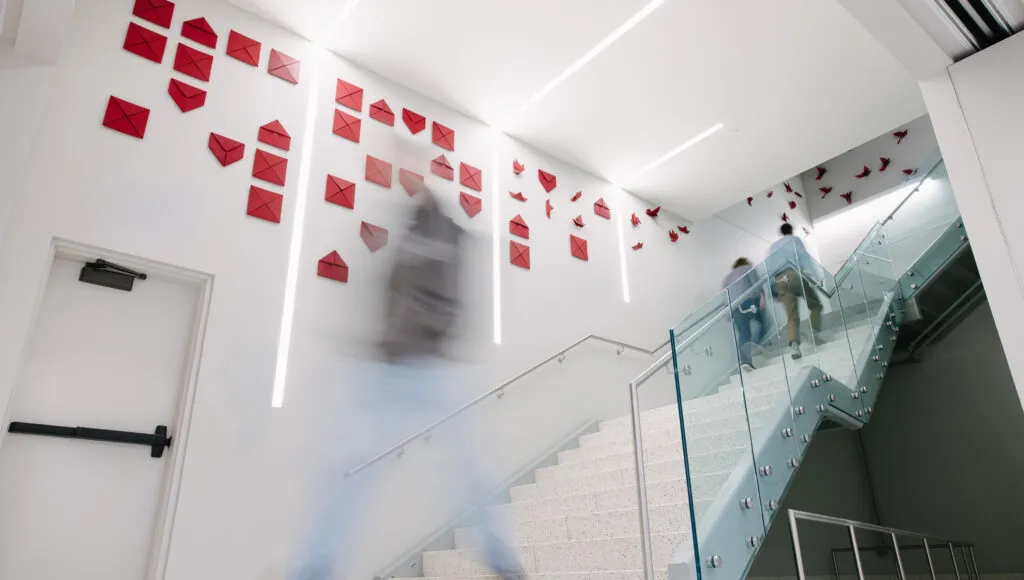 Dozens of origami birds placed on wall alongside staircase, birds slowly transition from flat paper into complex folded design.